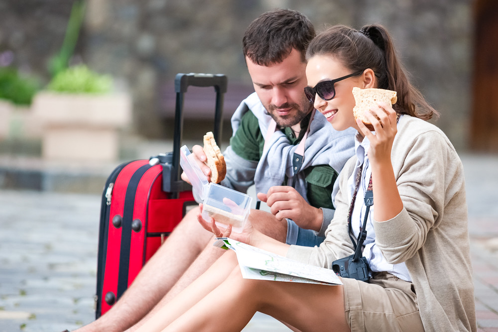 couple looking at map, eating sandwich, with red suitcase